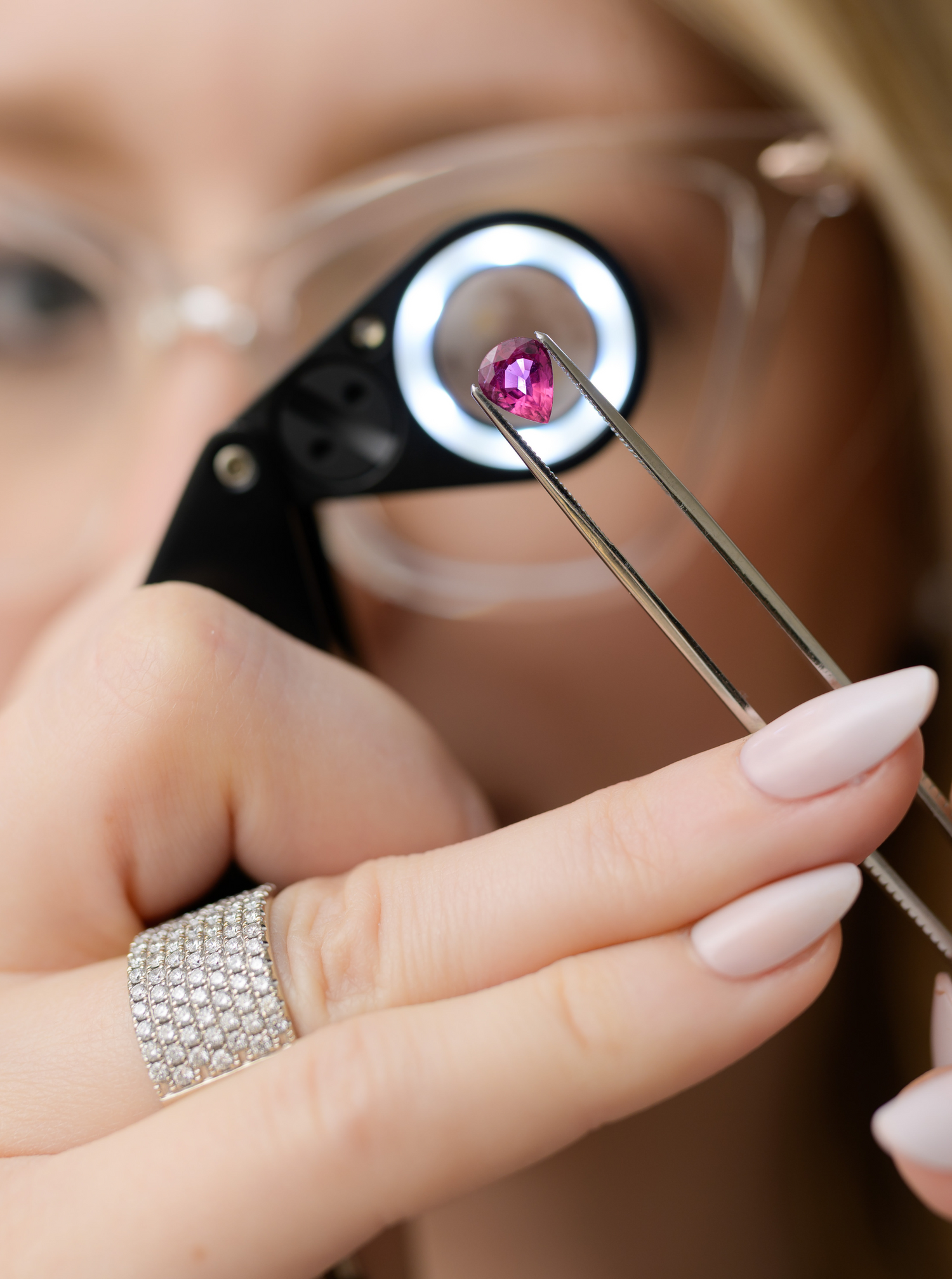 Expert jeweler inspecting a stunning pink gemstone with precision tools under a magnifying loupe. Shop fine jewelry at Devine Jeweler.